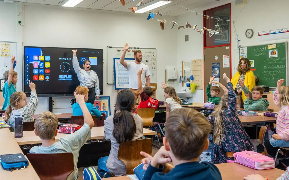 Streitkultur in der Grundschule – Projekt „Cool bleiben – fair streiten“ Grundschulklasse hebt die Hände, während zwei Theaterpädagogen und eine Lehrerin vorn im Klassenraum stehen