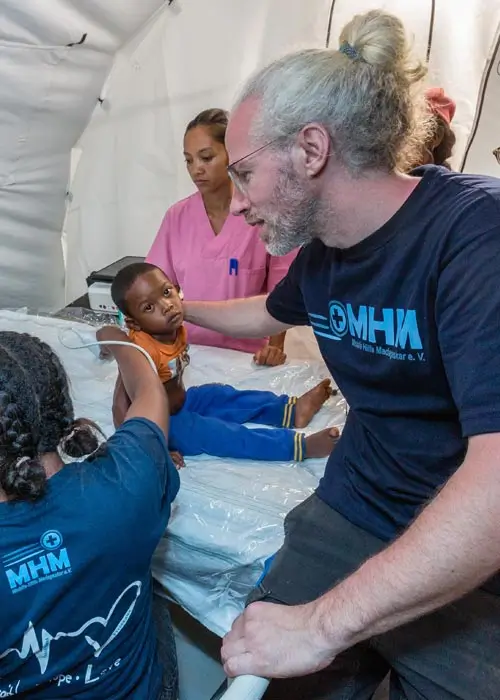 DEICHMANN-Stiftung | Madagascar Mobile Clinic – medizinisches Personal befähigen – Dr. Fabian Bendel unterstützt eine madagassische Ärztin, die sich mit dem Ultraschallgerät vertraut macht  Madagascar Mobile Clinic – Ärztin macht Ultraschall bei Kind, Arzt hält Jungen fest und blickt mit Ärztin auf Bildschirm