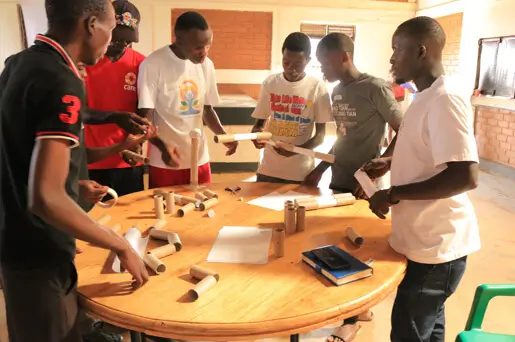 Deichmann Stiftung | Sozialunternehmen Uganda – Persönlichkeitsentwicklung und erfahrungsbasiertes Lernen Sozialunternehmen Uganda – mehrere Personen stehen an einem runden Tisch und schauen sich als Team gemeinsam Dokumente und Papierrollen an – erfahrungsbasiertes Lernen